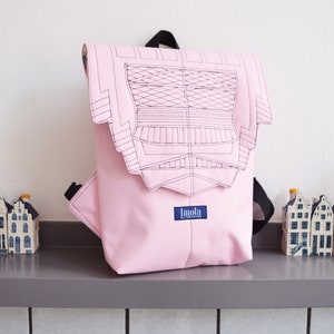 Puede incluir: Una mochila rosa con un gráfico en blanco y negro de un edificio en la solapa delantera. La mochila tiene una correa negra y una etiqueta azul que dice "Imola by coconino".