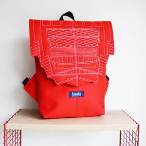 Puede incluir: Una mochila roja brillante con detalles de costura blancos en la solapa. La mochila tiene correas negras y una pequeña etiqueta azul con texto blanco. La bolsa está sobre una estantería de madera clara con un marco de metal rojo.