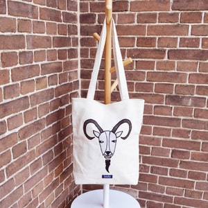 May include: A white tote bag with a black and white embroidered image of a goat's head. The bag has a blue label that says "Linda".
