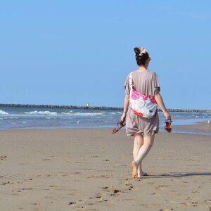 K&ouml;nnte beinhalten: Eine Person geht an einem sonnigen Tag an einem Sandstrand entlang und tr&auml;gt Sandalen. Sie tr&auml;gt ein kurzes, taupefarbenes Kleid und eine Umh&auml;ngetasche mit Blumenmuster. Im Hintergrund sind das Meer und ein klarer blauer Himmel zu sehen.