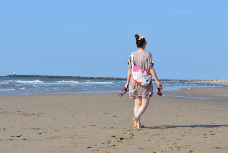 K&ouml;nnte beinhalten: Eine Person geht an einem sonnigen Tag an einem Sandstrand entlang und tr&auml;gt Sandalen. Sie tr&auml;gt ein kurzes, taupefarbenes Kleid und eine Umh&auml;ngetasche mit Blumenmuster. Im Hintergrund sind das Meer und ein klarer blauer Himmel zu sehen.