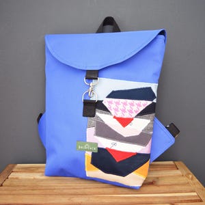 Puede incluir: Una mochila azul con cierre de solapa y un bolsillo frontal con un diseño de patchwork de varios colores y patrones. La mochila tiene correas negras y un cierre plateado. La marca "boldpeach" es visible en una pequeña etiqueta verde.