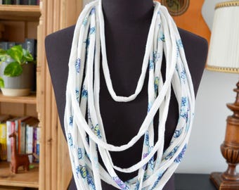 Pode incluir: Colar de tecido branco com um padrão floral em azul e roxo. O colar é exibido em um manequim preto. O fundo inclui uma estante, uma guitarra e uma lâmpada.