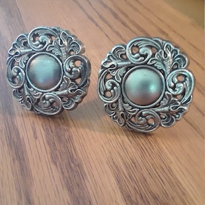 May include: Two silver-toned metal drawer pulls with ornate floral designs. The pulls have a round, slightly raised center.