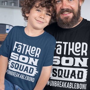 Op de afbeelding: Twee bijpassende T-shirts, een donkerblauw en een zwart, met de tekst "Father Son Squad" en "#Unbreakablebond" in wit. De shirts worden gedragen door een vader en zijn zoon.