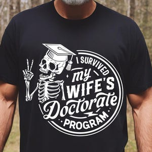 Puede incluir: Camiseta negra con un gráfico blanco de un esqueleto con un gorro de graduación y haciendo un signo de paz. El gráfico está dentro de un círculo con el texto "I Survived My Wife's Doctorate Program".