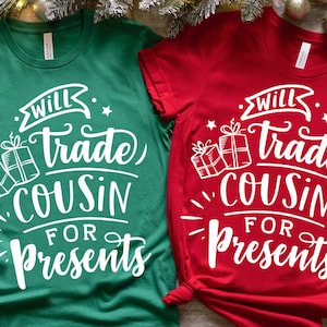 May include: Two green and red t-shirts with white text that reads "Will trade cousin for presents". The shirts are laid out on a wooden surface with Christmas decorations.