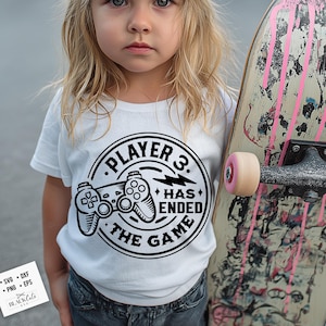 May include: A white t-shirt with a black graphic that reads "PLAYER 3. HAS ENDED THE GAME" with a game controller design. The child is holding a skateboard with pink accents.