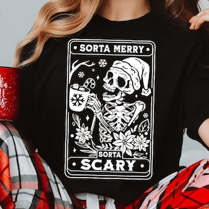 May include: A black t-shirt with a white graphic of a skeleton wearing a Santa hat and holding a mug of hot chocolate. The graphic is surrounded by a white border and the text "Sorta Merry" and "Sorta Scary" in white.