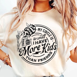 May include: Cream-colored t-shirt with a black graphic. The graphic features a skeleton with a bun and the text "MY CIRCLE IS so small I HAVE More Kids THAN FRIENDS". The skeleton is giving a peace sign.