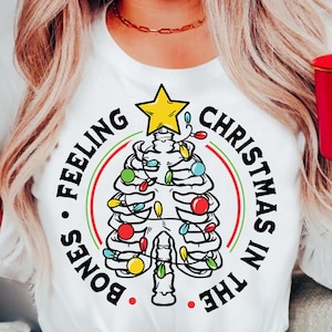 Op de afbeelding: Een wit longsleeve shirt met een zwarte cirkelvormige afbeelding van een skelet met kerstverlichting. De tekst "Bones Feeling Christmas in the" staat rond de cirkel.