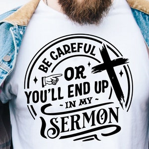 以下が含まれることがあります： 白いTシャツに、黒い円形のデザインが施されています。デザインには「Be Careful Or You'll End Up In My Sermon」という文字と、十字架と指さす手が描かれています。