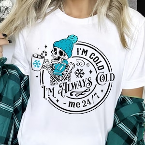May include: A white t-shirt with a black and white graphic design of a skeleton wearing a blue hat and scarf holding a mug of hot chocolate. The graphic design includes the text "I'm Cold I'm Always Cold - me 24/7".