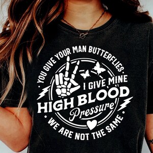 May include: Black t-shirt with a white circle graphic featuring a skeleton hand making a peace sign, butterflies, and the text "You give your man butterflies. I give mine high blood pressure. We are not the same."
