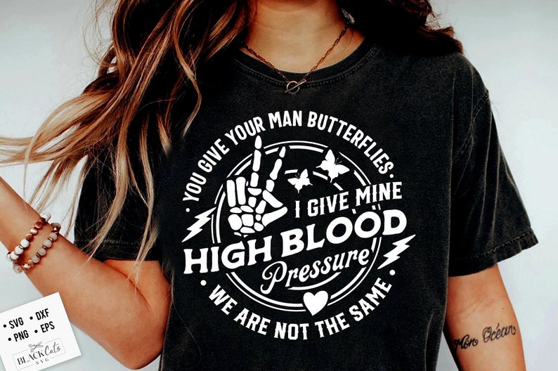 May include: Black t-shirt with a white circle graphic featuring a skeleton hand making a peace sign, butterflies, and the text "You give your man butterflies. I give mine high blood pressure. We are not the same."