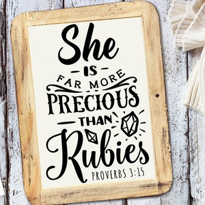 Puede incluir: Cartel enmarcado en madera con el texto "She is far more precious than rubies Proverbs 3:15" en escritura negra. El cartel tiene un fondo crema y un marco de madera marrón claro. Una taza de café y un suéter blanco están en el fondo.