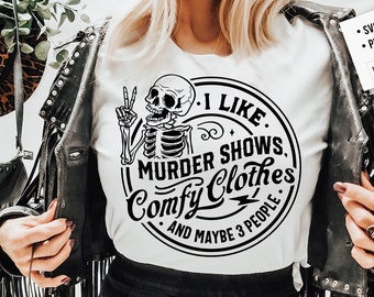 May include: A white t-shirt with a black and white graphic of a skeleton holding a peace sign. The graphic is inside a circle with the text "I Like Murder Shows, Comfy Clothes, And Maybe 3 People".