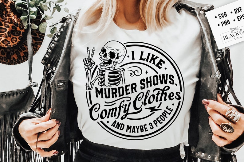 May include: A white t-shirt with a black and white graphic of a skeleton holding a peace sign. The graphic is inside a circle with the text "I Like Murder Shows, Comfy Clothes, And Maybe 3 People".
