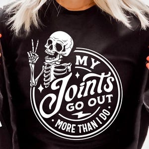 May include: Black sweatshirt featuring a white skeleton graphic giving a peace sign. The text "MY Joints GO OUT MORE THAN I DO" is in a circular design.