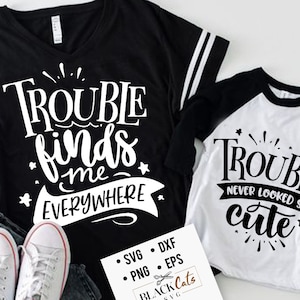 Puede incluir: Dos camisetas blancas y negras con un diseño gráfico blanco. La camiseta más grande tiene el texto "Trouble finds me everywhere". La camiseta más pequeña tiene el texto "Trouble never looked so cute".