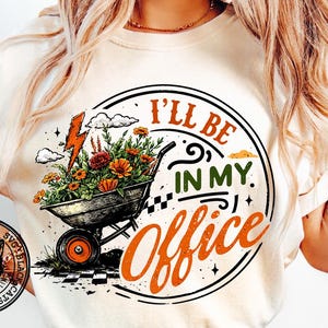 以下が含まれることがあります： クリーム色のTシャツ。円形のグラフィックには、花でいっぱいの手押し車、稲妻、そして「I'll Be In My Office」というテキストが遊び心のあるフォントで描かれています。黒猫のロゴが見えます。