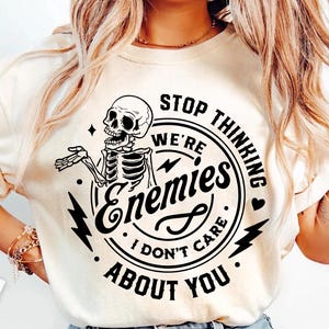 May include: Cream-colored t-shirt with a black graphic featuring a skeleton and the text "STOP THINKING WE'RE ENEMIES I DON'T CARE ABOUT YOU" in a circular design. The design includes lightning bolts and stars.