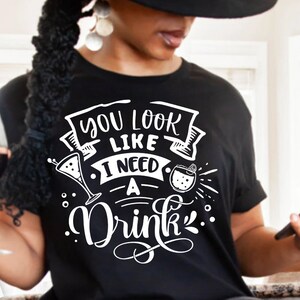 May include: A black t-shirt with white text that reads "You look like I need a drink". The text is in a fun, handwritten style and includes illustrations of cocktails.