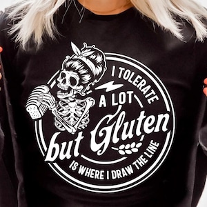 以下が含まれることがあります： 黒いTシャツに白黒のグラフィックデザイン。デザインは、パンを持った骸骨が「私はたくさん我慢するけど、グルテンは私の限界」というテキストと共に描かれている。
