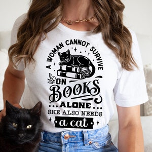 May include: A white t-shirt with a black graphic that reads "A woman cannot survive on books alone... she also needs a cat." The graphic features a black cat sitting on a stack of books.