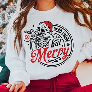 May include: White sweatshirt with a graphic of a skeleton wearing a Santa hat, holding a mug with a snowflake design and a candy cane. The text reads "DEAD INSIDE but Merry" in a circular design. A festive holiday theme.