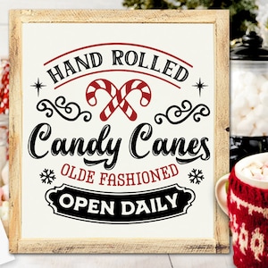 May include: A wooden framed sign with the text "HAND ROLLED Candy Canes OLDE FASHIONED OPEN DAILY" in black and red. The sign features candy cane graphics and snowflake accents. The background includes a mug of hot chocolate and other festive decorations.
