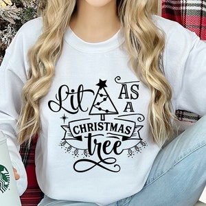 以下が含まれることがあります： 黒い文字で「Lit as a Christmas tree」と書かれた白いスウェットシャツ。テキストは、ライトとバナーが付いたクリスマスツリーで囲まれています。