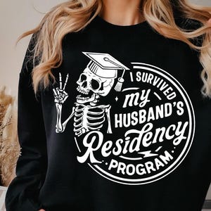 May include: Black sweatshirt with a white graphic featuring a skeleton wearing a graduation cap and giving a peace sign. The text reads "I SURVIVED my HUSBAND'S Residency PROGRAM."