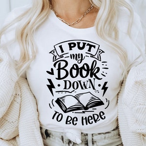Könnte beinhalten: Ein weißes T-Shirt mit schwarzem Text, der "Ich habe mein Buch abgelegt, um hier zu sein" lautet. Der Text ist in einer stilisierten Schriftart gehalten und enthält eine Zeichnung eines offenen Buches mit Blitzen.