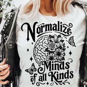 May include: White t-shirt with the words "Normalize Minds of all Kinds" in black script. The design includes a brain, flowers, and butterflies. The person is wearing a black leather jacket.