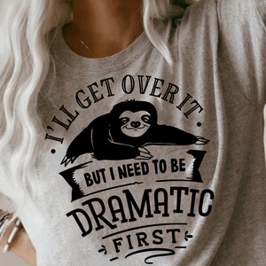 Puede incluir: Una camiseta gris con un gráfico negro de un perezoso. El gráfico dice "Lo superaré, pero necesito ser dramático primero."