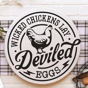Può includere: Un cartello rotondo in legno bianco con testo nero che recita "Wicked Chickens Lay Deviled Eggs". Al centro del cartello è presente una silhouette nera di una gallina.