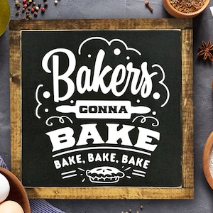 Könnte beinhalten: Ein schwarzes Schild aus Schiefer mit weißem Text, der "Bakers Gonna Bake" und "Bake, Bake, Bake" lautet. Das Schild ist in Holz gerahmt und hat eine kleine Tortengrafik unter dem Text.