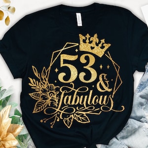 Op de afbeelding: Een zwart T-shirt met een gouden glitterontwerp met de tekst "53 & Fabulous" met een kroon en bloemmotieven.