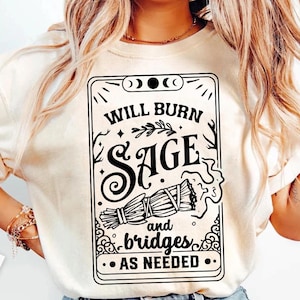 Könnte beinhalten: Cremefarbenes T-Shirt mit schwarzem Grafikdesign. Das Design zeigt den Text "WILL BURN SAGE and bridges AS NEEDED" in einem dekorativen Rahmen, zusammen mit einer Illustration eines Salbeibündels. Das Shirt ist ein Freizeitartikel.