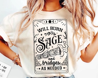 May include: Cream-colored t-shirt with a black graphic design. The design features the text "WILL BURN SAGE and bridges AS NEEDED" within a decorative frame, along with an illustration of a sage bundle. The shirt is a casual wear item.
