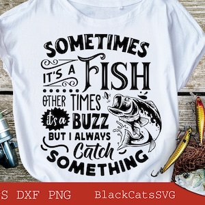 May include: White t-shirt with a black graphic that reads "Sometimes it's a fish, other times it's a buzz, but I always catch something." Fishing lures, a fishing reel, and a fish net are arranged around the shirt.