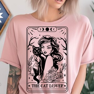 May include: A light pink t-shirt featuring a black graphic of a woman holding a black cat, framed by a decorative border with the text "THE CAT LOVER". The design includes floral and celestial elements.