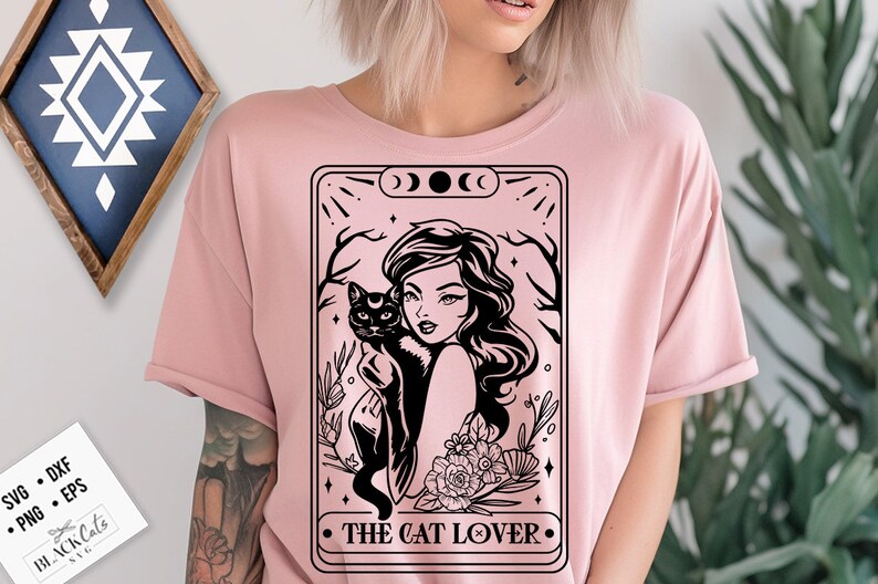 May include: A light pink t-shirt featuring a black graphic of a woman holding a black cat, framed by a decorative border with the text "THE CAT LOVER". The design includes floral and celestial elements.