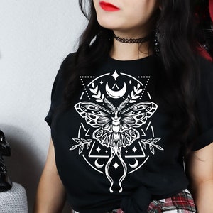 Puede incluir: Una camiseta negra con un diseño gráfico blanco de una polilla con formas geométricas y lunas crecientes. El diseño está centrado en la camiseta.