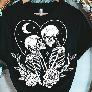 May include: Black t-shirt featuring a white graphic of two skeletons embracing within a heart, adorned with flowers and a crescent moon. The text "Black Cats SVG" is at the bottom of the image.