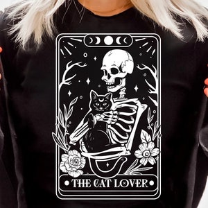 Puede incluir: Una ilustraci&oacute;n en blanco y negro de un esqueleto sosteniendo un gato negro. La imagen est&aacute; en el estilo de una carta de tarot y tiene el texto "THE CAT LOVER." en la parte inferior.
