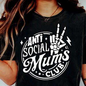 Op de afbeelding: Zwart T-shirt met een witte cirkelvormige afbeelding met de tekst "Anti Social Mums Club" en een skelet hand die een peace-teken maakt.