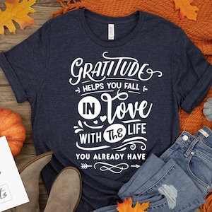 以下が含まれることがあります： 「Gratitude helps you fall in love with the life you already have」の白文字がプリントされたネイビーブルーのTシャツ。ジーンズ、ブーツ、カボチャ、秋の葉が写っています。