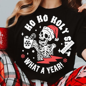 Può includere: T-shirt nero con uno scheletro con un cappello da Babbo Natale, che tiene in mano una tazza con marshmallow e un bastoncino di zucchero. La maglietta dice "HO HO HOLY S**T WHAT A YEAR!" in testo bianco. Una tazza natalizia rossa e bianca è tenuta nella mano sinistra.